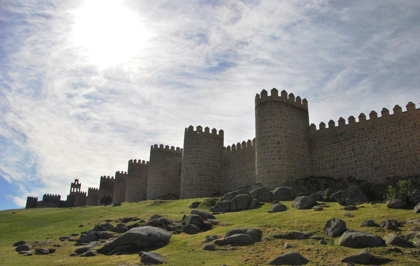 The medieval walls and towers of Ávila seen from below, with granite boulders on the slope and the sun behind the clouds